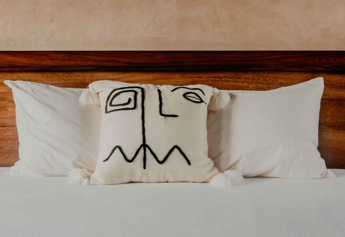 Hand-loomed pillow and embroidered by skilled artisans with a geometric face inspired by the Cubism movement stitched on a cream ground finished with white pom poms.