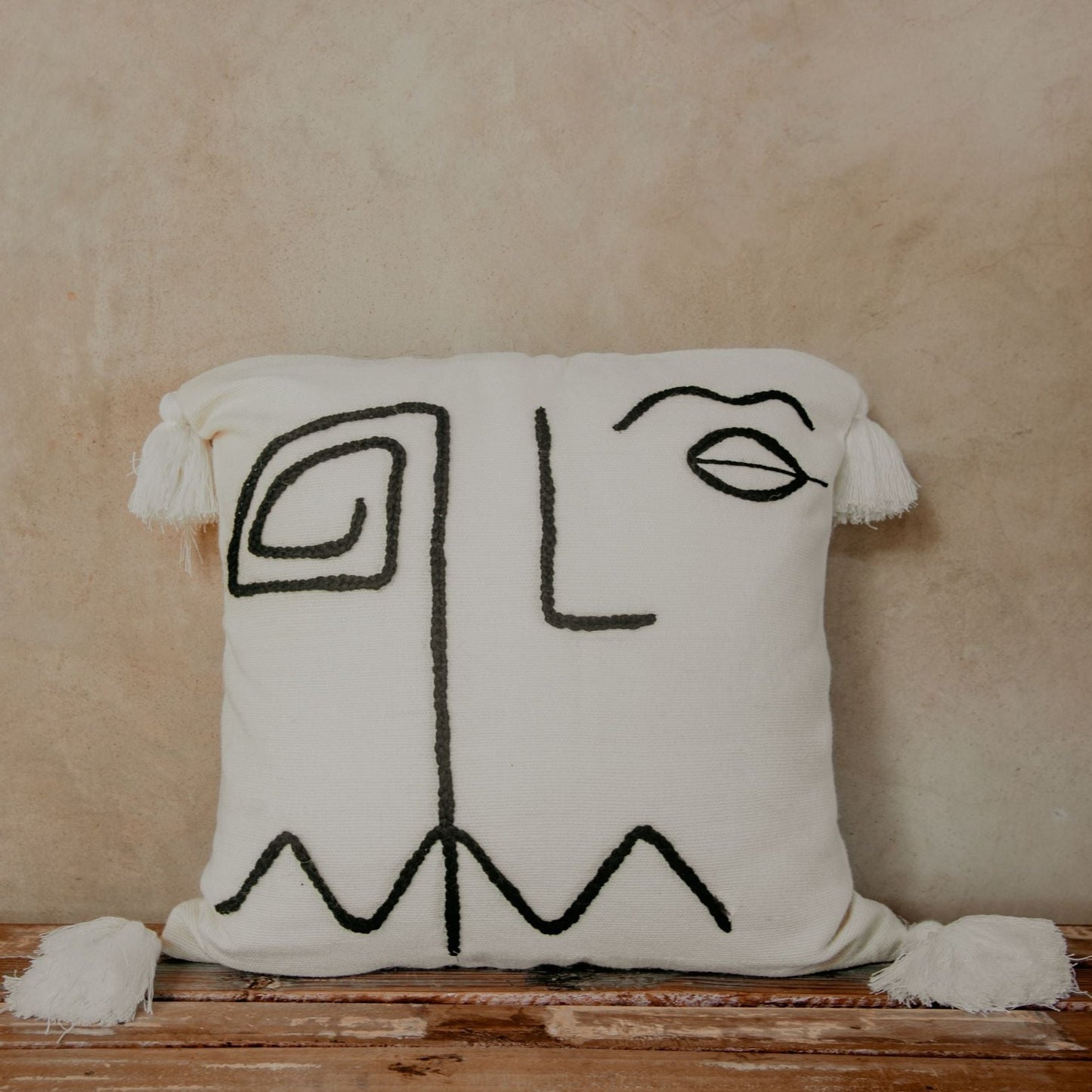 Hand-loomed pillow and embroidered by skilled artisans with a geometric face inspired by the Cubism movement stitched on a cream ground finished with white pom poms.